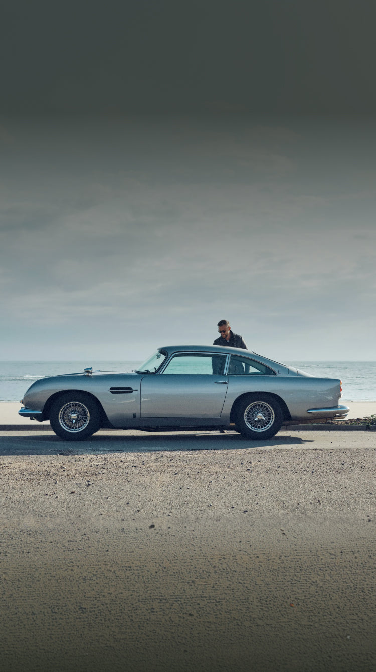 Silver classic car on a beach with a person standing next to it