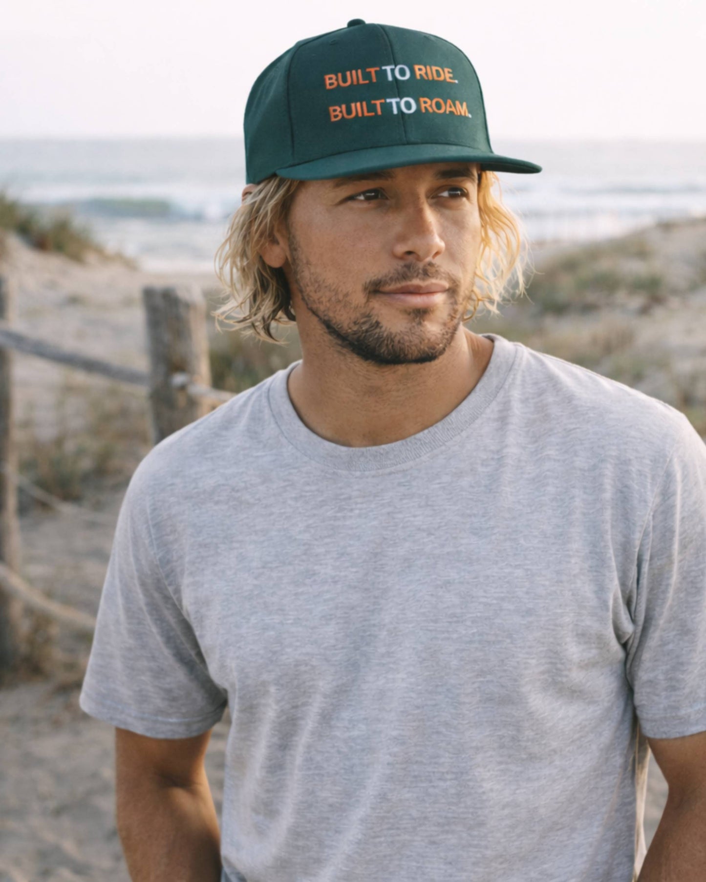 Man wearing a cap with 'Built to Ride, Built to Roam' text on a beach.