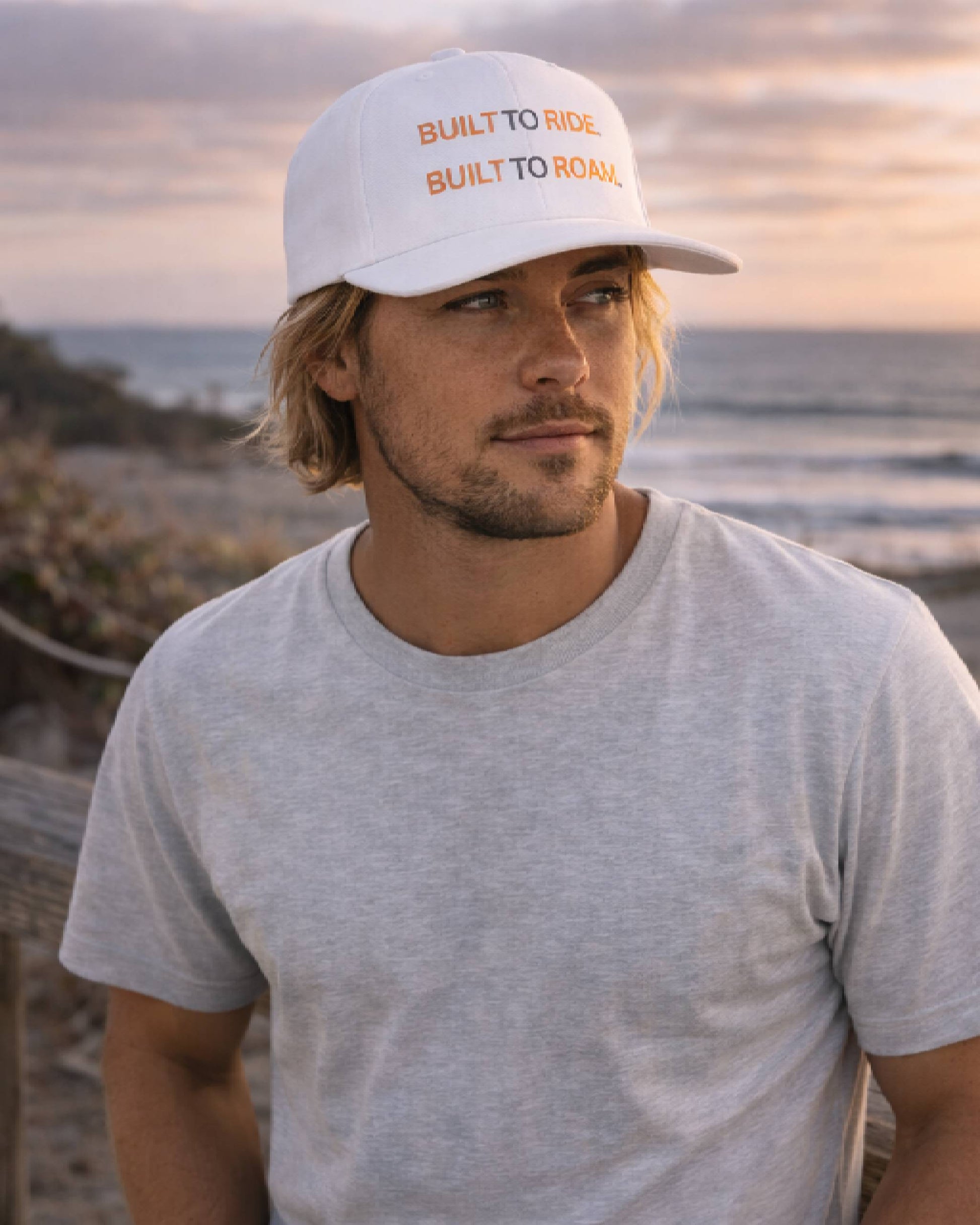 Man wearing a white cap with text, standing by the ocean.