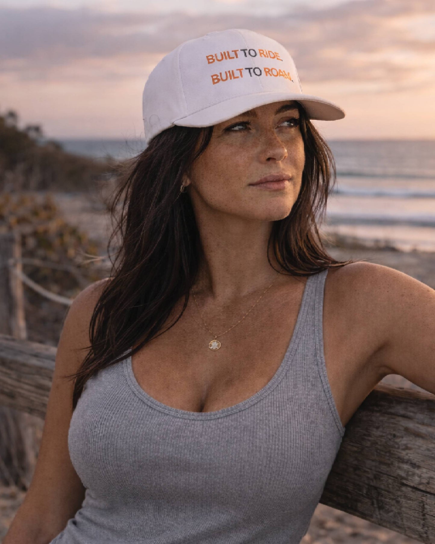 Woman wearing a white cap with text, standing on a beach at sunset.