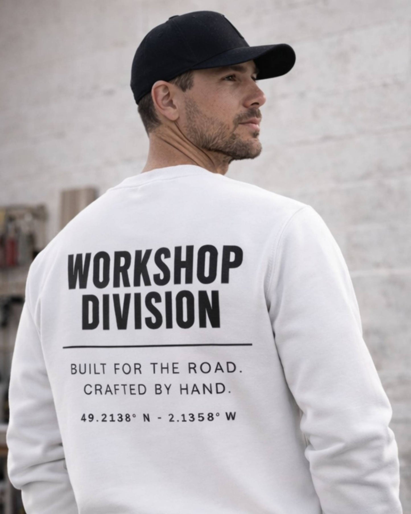 Man wearing a white sweatshirt with 'Workshop Division' text on the back, standing against a light gray wall.