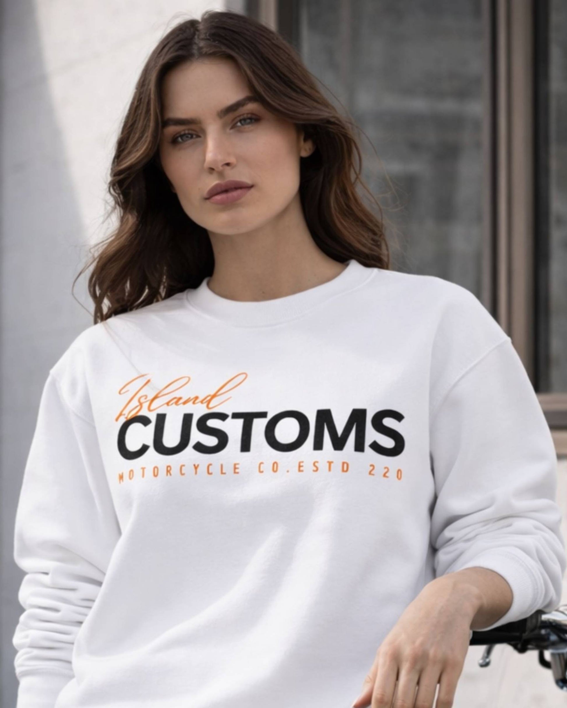 Woman wearing a white sweatshirt with 'Island Customs' text, sitting outdoors.