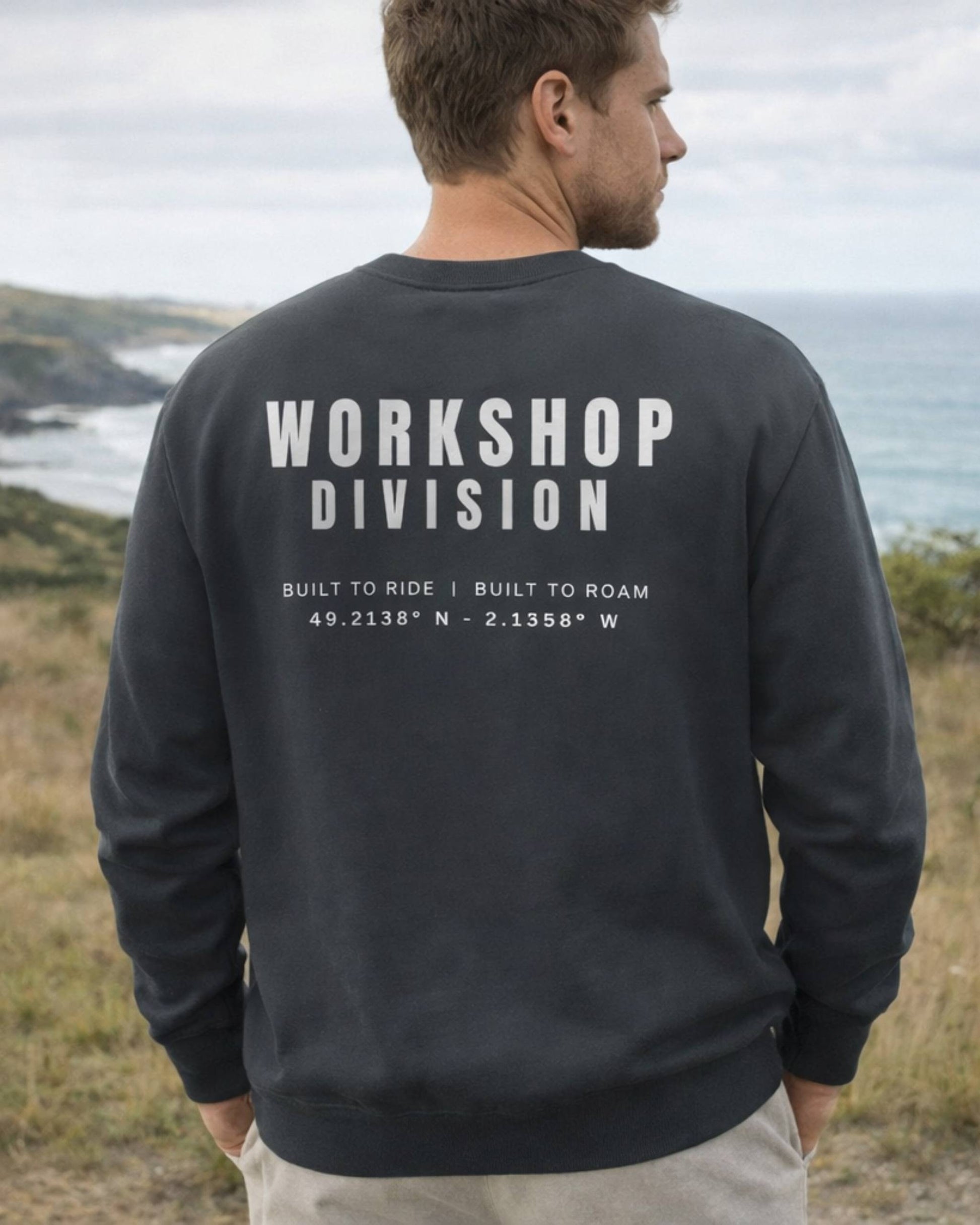 Man wearing a black sweatshirt with 'Workshop Division' text outdoors by the ocean.