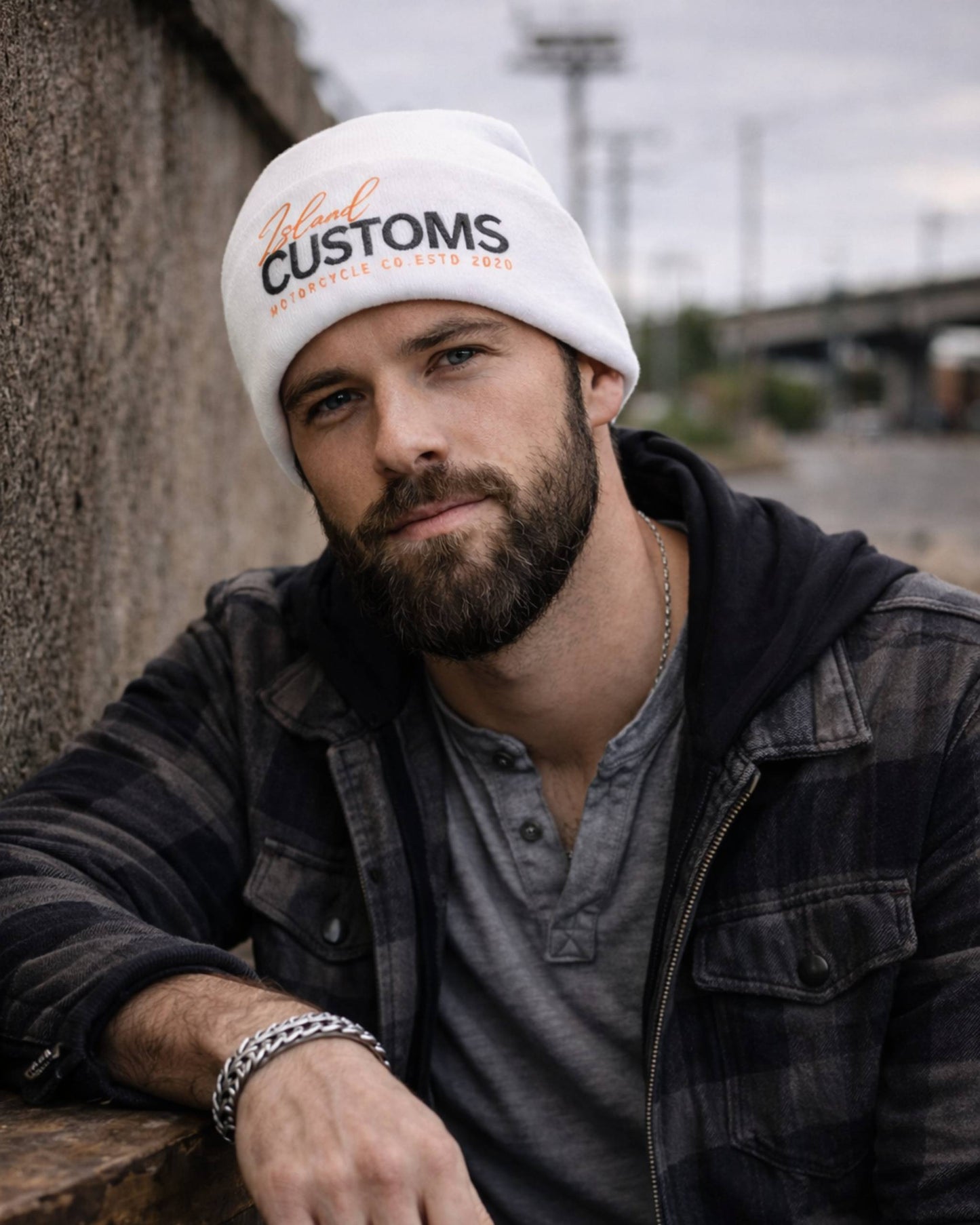 Man wearing a white beanie with 'Customs' text, leaning against a wall outdoors.