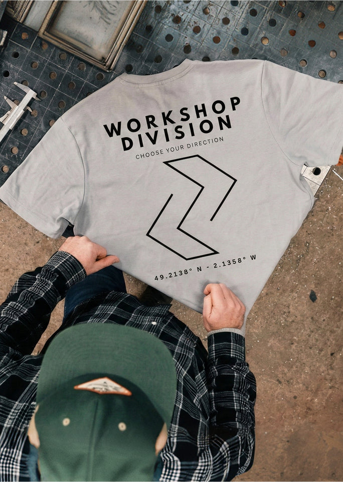 Gray t-shirt with 'Workshop Division' design held by a person wearing a plaid shirt and green cap.