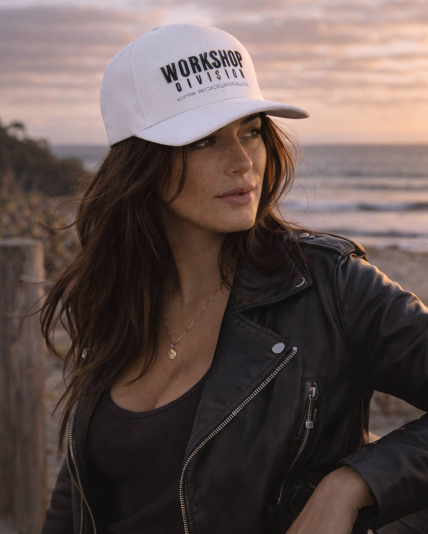 Woman wearing a white cap with 'Workshop Division' text on a beach at sunset.