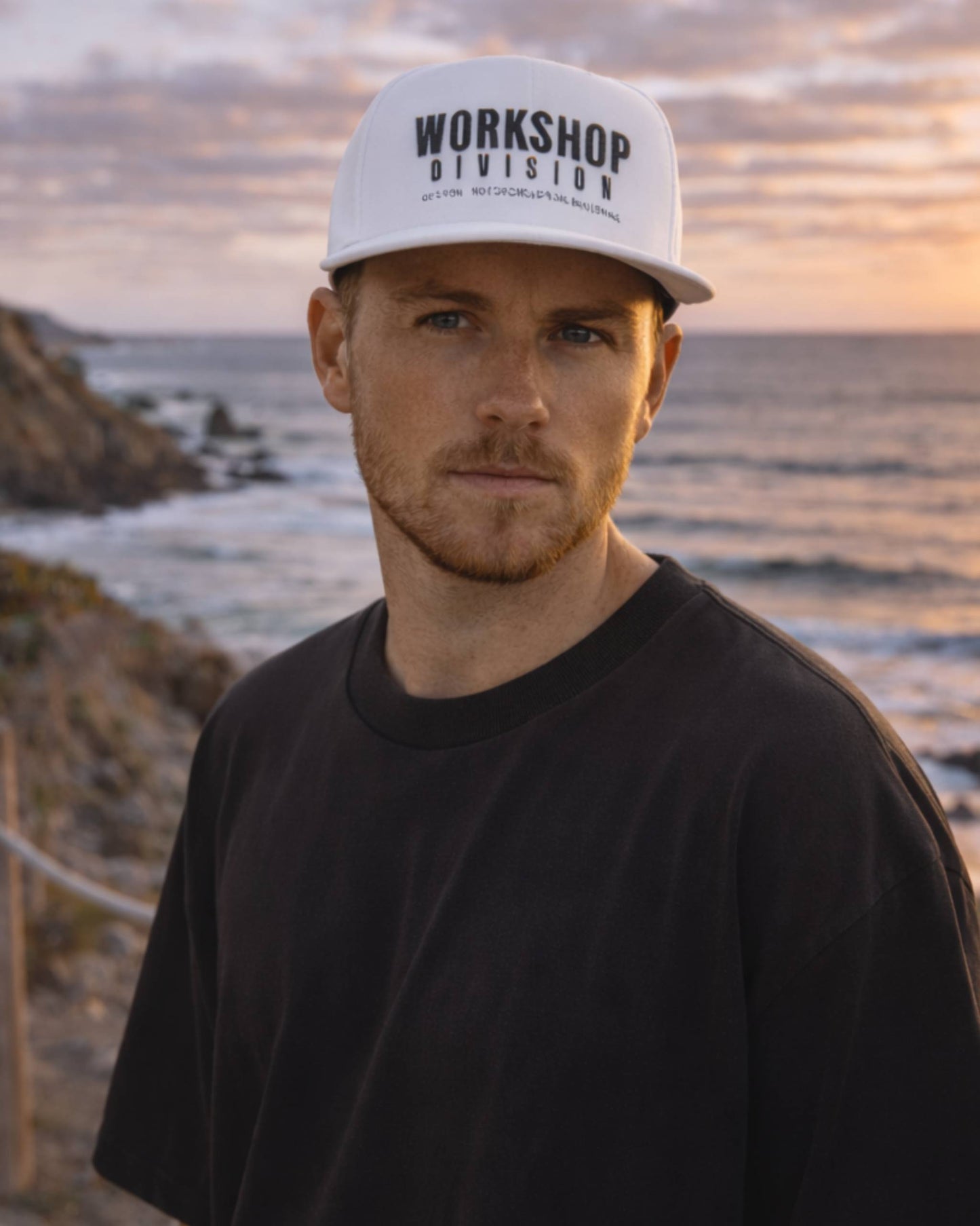Man wearing a 'Workshop Division' cap with a beach and sunset in the background