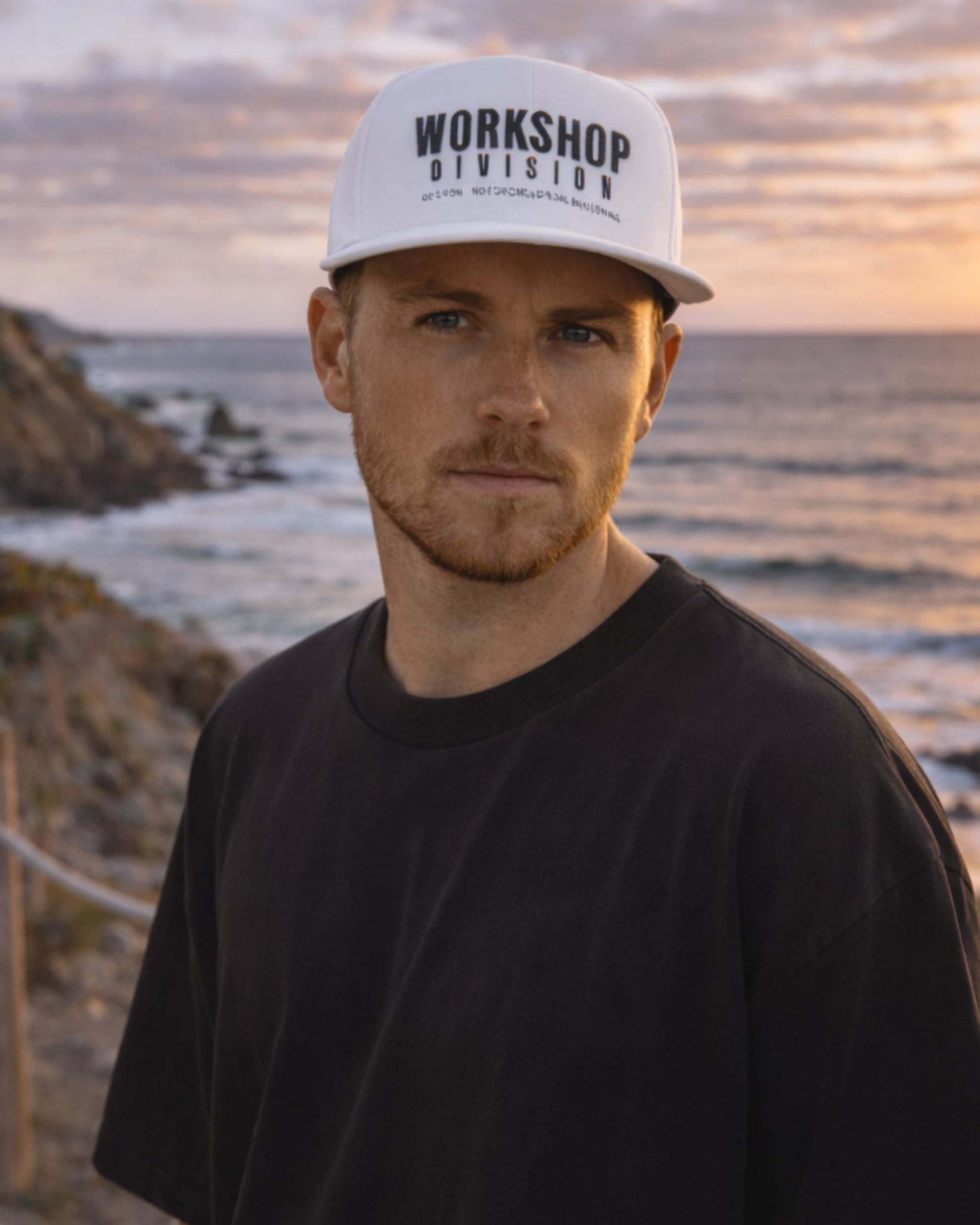 Man wearing a 'Workshop Division' cap with a beach and sunset in the background