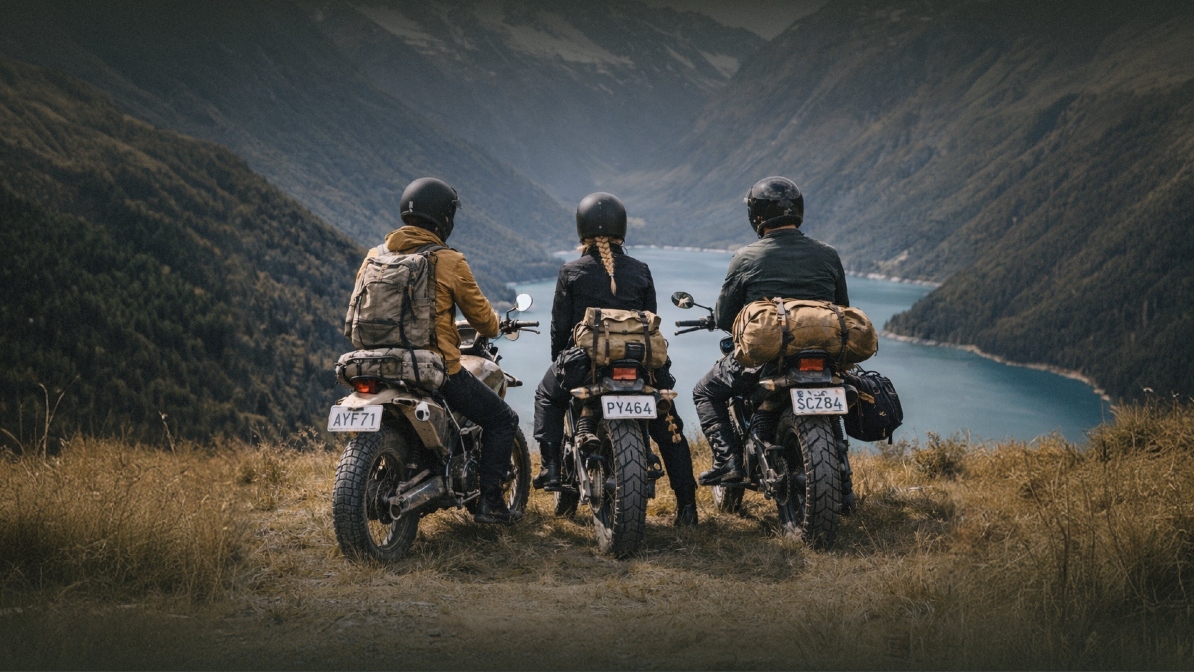 Three people on motorcycles with gear, overlooking a scenic valley with a lake.