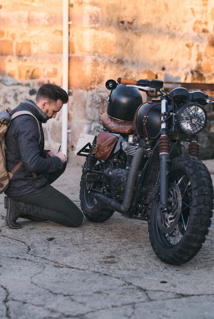 Man inspecting a custom motorcycle against a stone wall.