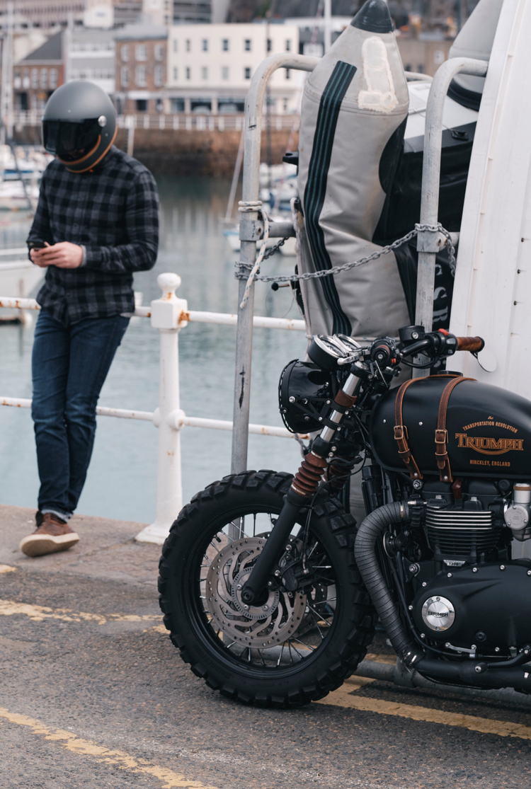 Person with a helmet standing next to a Triumph motorcycle by a waterfront.