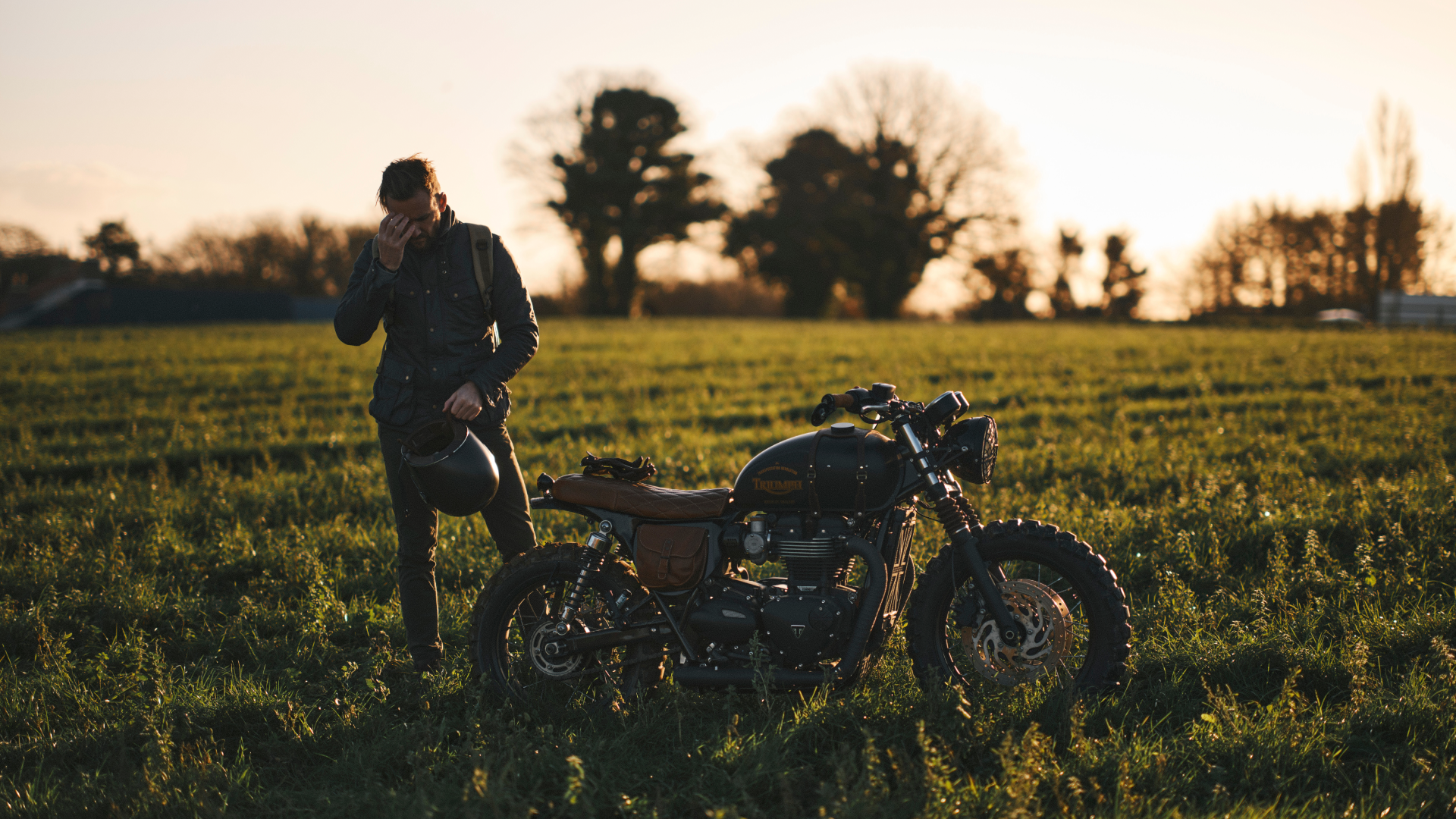 Person standing next to a motorcycle in a field at sunset