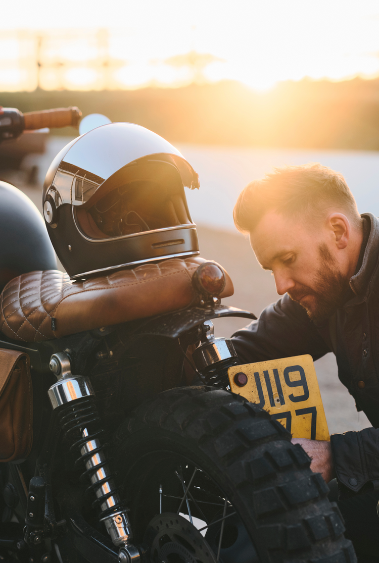 Man inspecting a motorcycle with a helmet on the seat, sunset in the background