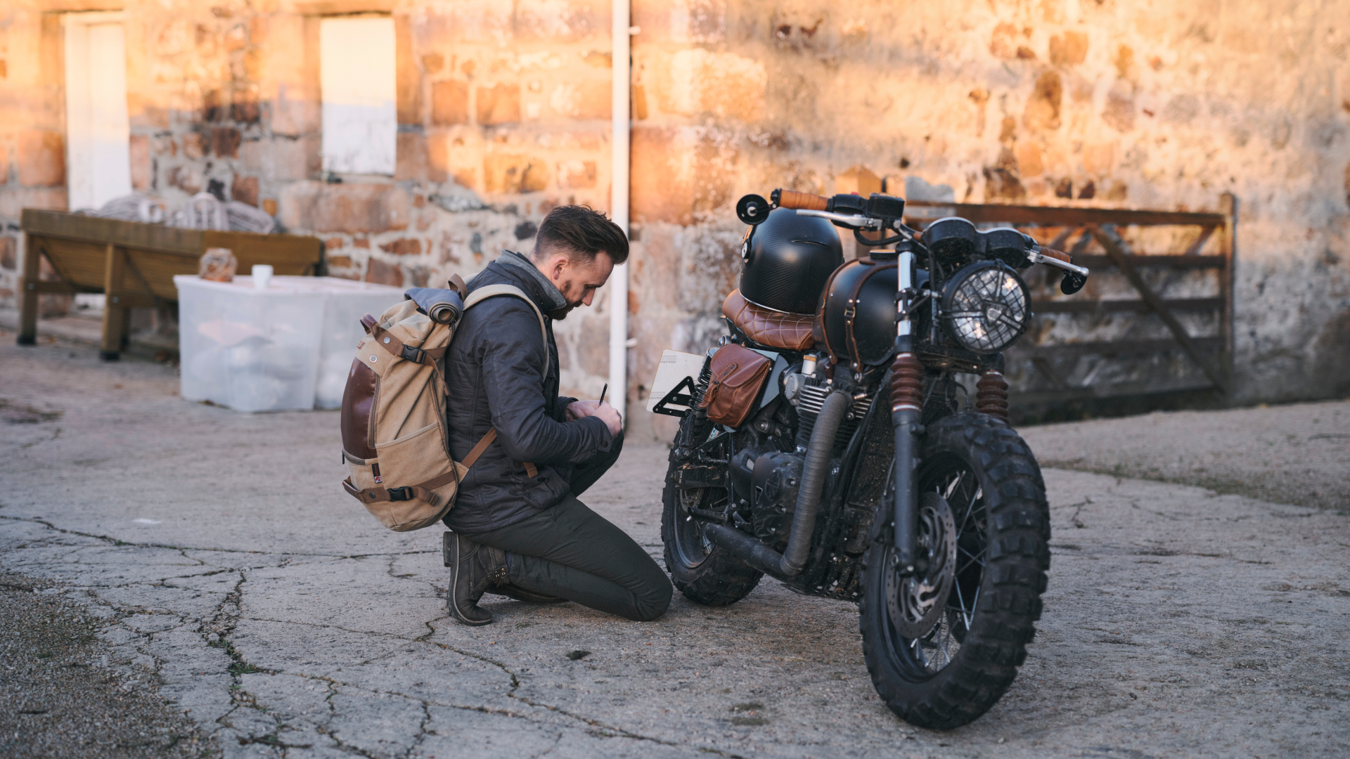 Man kneeling next to a motorcycle in an outdoor setting with stone walls.