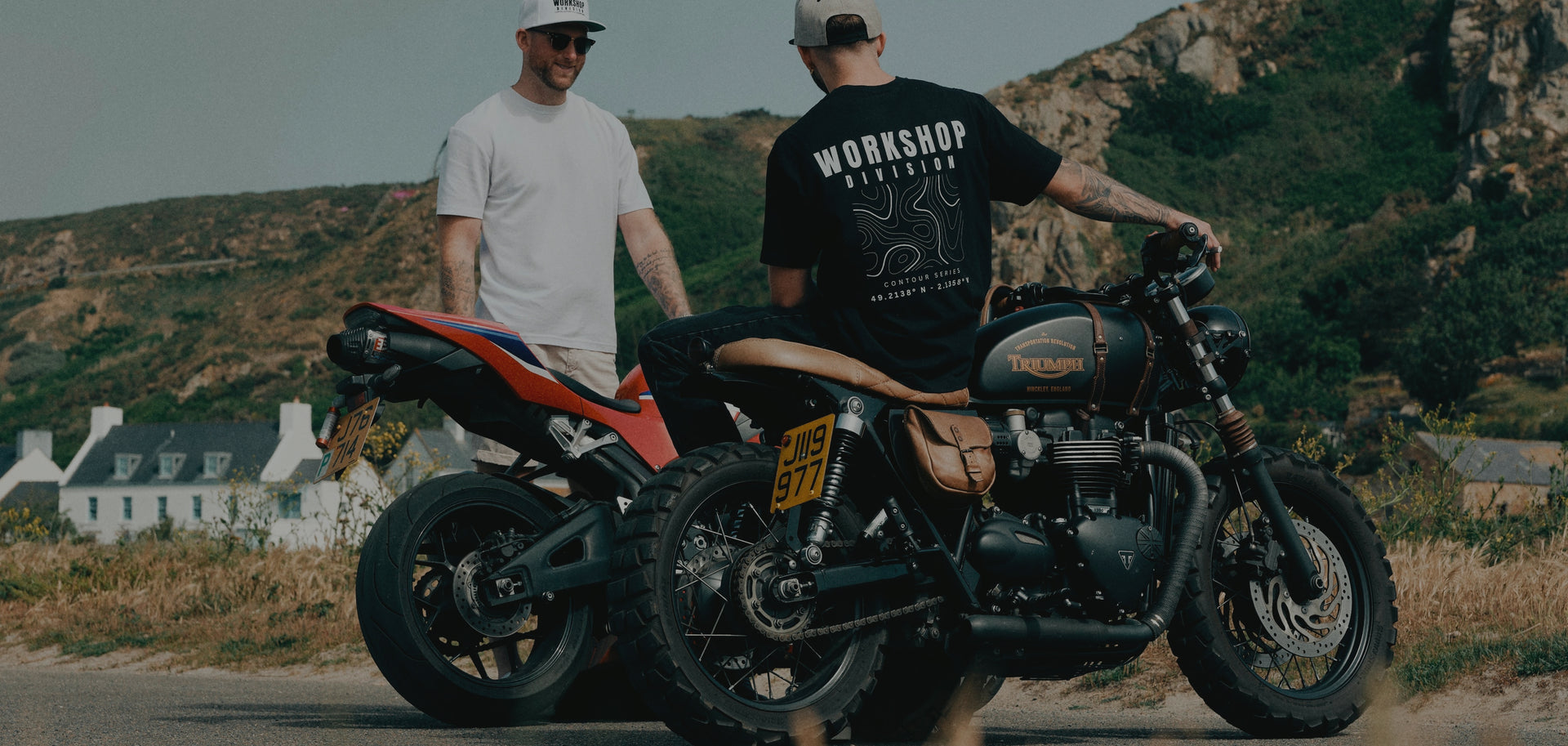 Two men standing next to a motorcycle on a scenic road with houses and greenery in the background.
