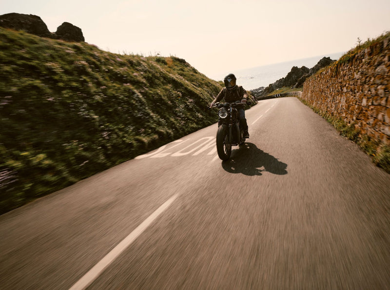 Person riding a motorcycle on a winding road with scenic hills and stone wall.